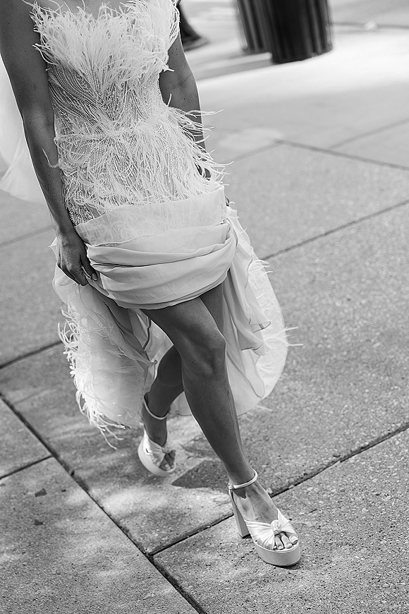 Wedding dress close-up showing feather wedding dress trim, beaded bodice, high slit and ankle-strap platform heels on a city sidewalk