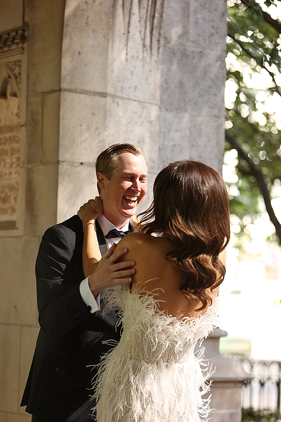 Wedding couple portrait of bride and groom laughing in a close embrace, her strapless feather-trim gown beside his tuxedo under a stone archway