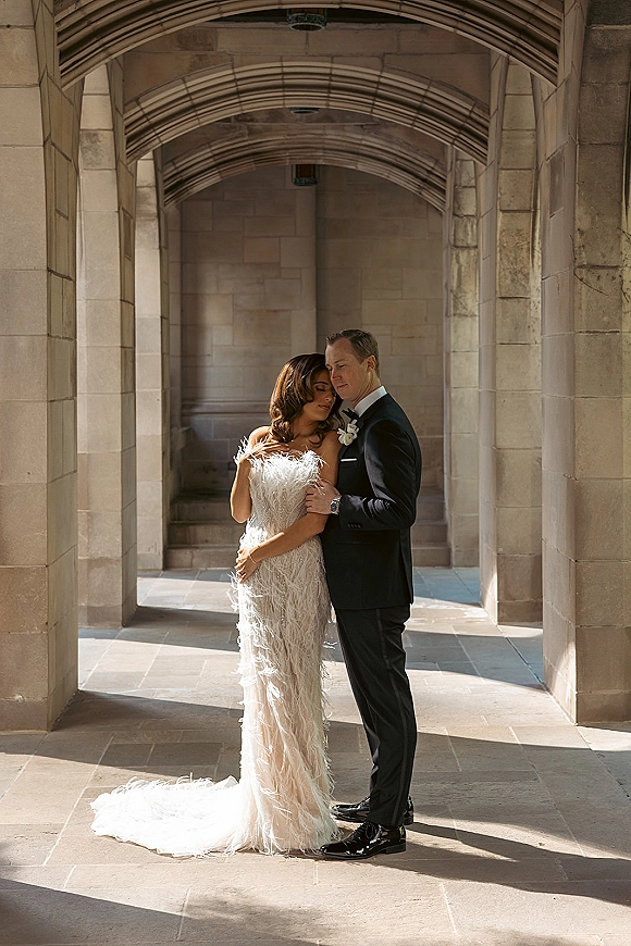 Couple portrait of bride and groom embrace under a sunlit stone archway corridor, her strapless feather wedding dress train flowing beside him