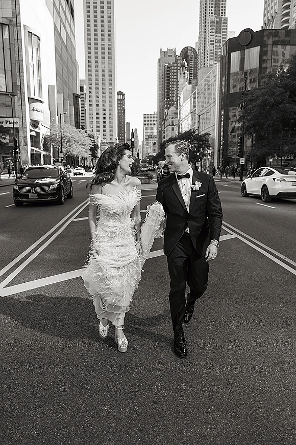Couple portrait of bride and groom walking holding hands on a city street, her strapless feather wedding dress and his tuxedo with bow tie