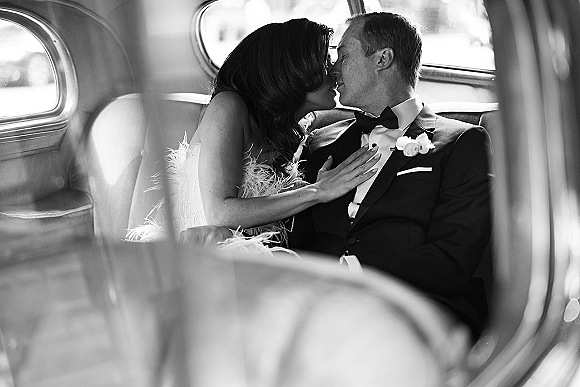 Wedding kiss portrait of bride and groom kissing in a vintage car, her feather-trim dress against his black tuxedo in window light