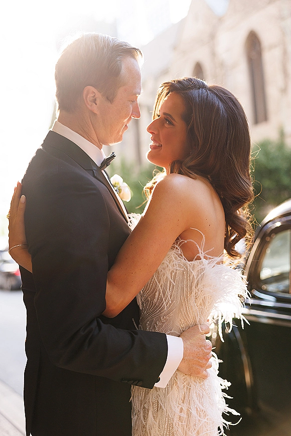 Couple portrait of bride and groom embrace beside a classic car, backlit by sun flare on a street with greenery and building facade