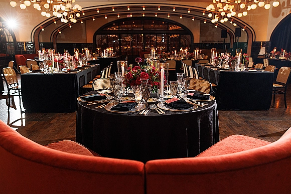 Reception tablescape with black tablecloth wedding style, gold flatware, black napkins, candles and red rose centerpiece under string lights in a hall