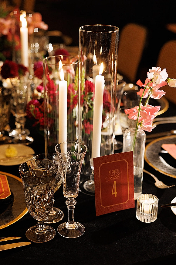 Reception tablescape with wedding table centerpiece of pink flowers, taper candles and glass cylinders on black tablecloth in dim lighting