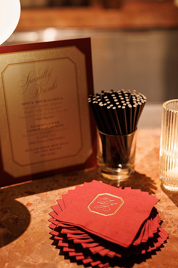Wedding cocktail napkins with custom monogram napkins in red and gold foil, paired with black stirrers and candlelight on a stone tabletop
