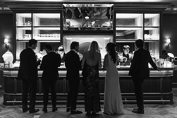 Wedding reception bar scene with tuxedoed guests and an evening gown ordering cocktails from a bartender at a mirrored hotel bar interior