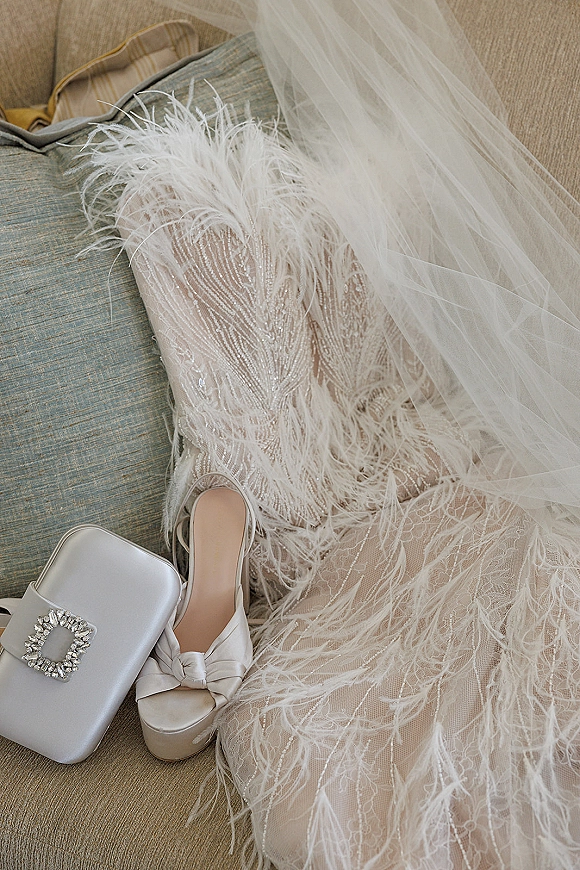 Wedding dress detail with feather trim and beaded lace bodice, styled with veil, satin platform heels and white clutch on sofa