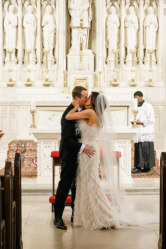 Wedding kiss at a church wedding kiss with the bride’s long veil and groom in tuxedo by gold candlesticks and red kneelers at the altar