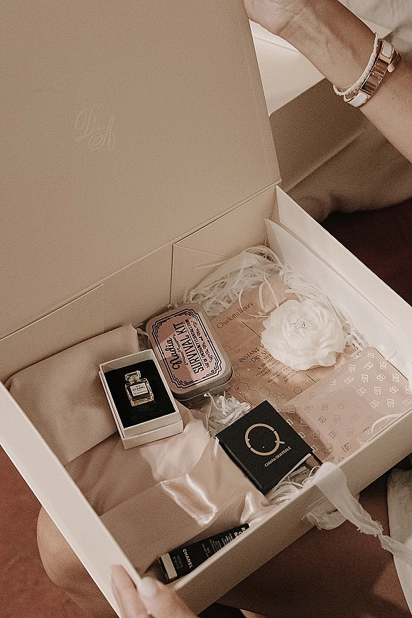 Wedding gift box with tissue paper and shredded filler, holding perfume, candle tin, satin pouch, watch, and a white flower in hands