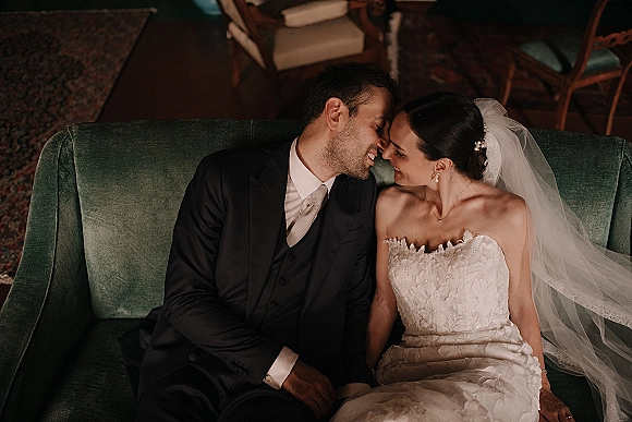 Couple portrait of bride and groom on couch, nose to nose on a green velvet sofa, her veil draped over shoulder in an indoor lounge