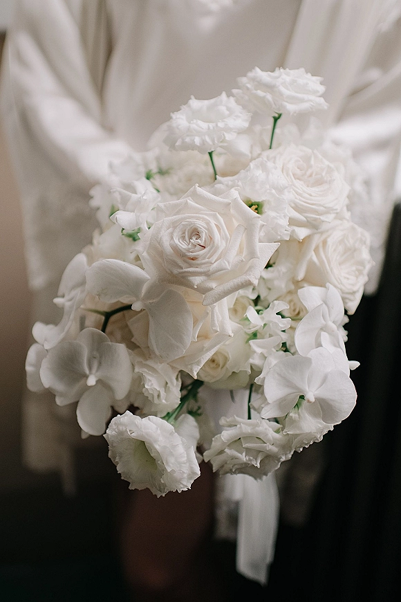 Bridal bouquet of white roses and orchids tied with a ribbon, held against a neutral indoor backdrop during getting ready