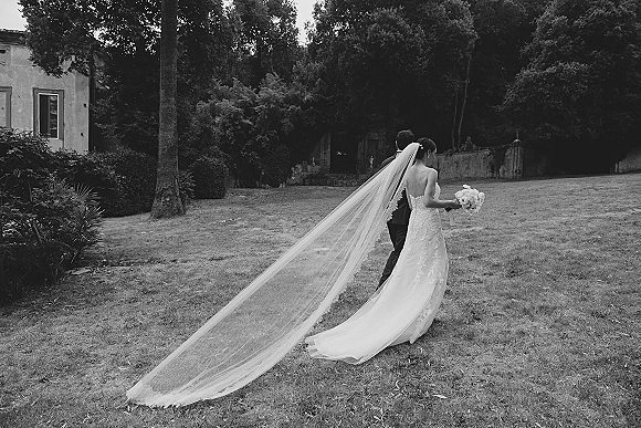 Bride and groom walking away, long cathedral veil trailing behind her strapless lace dress on a grassy villa garden lawn