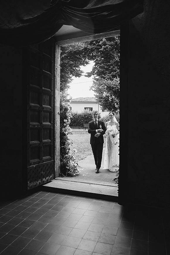 Wedding processional as bride walking down aisle with bouquet and veil, escorted by father through open wooden doors with rose petals and floral archway