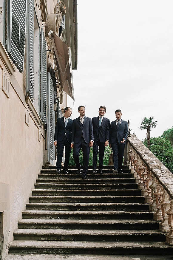 Groomsmen portrait of the groom with groomsmen in dark suits and ties standing on a stone staircase by a villa facade under overcast sky