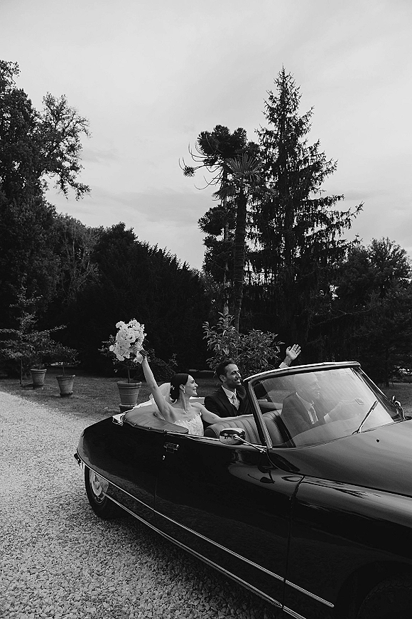 Wedding getaway car with newlyweds in a black classic convertible, bride raising bouquet as they ride down a gravel garden driveway