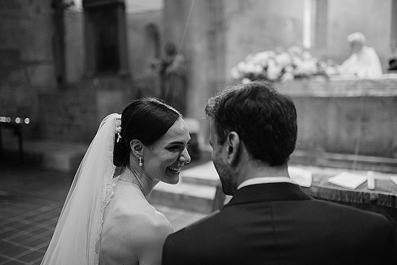 Ceremony moment in a black and white wedding photo as the bride smiles at the groom at the church altar, veil trailing by candlelit flowers