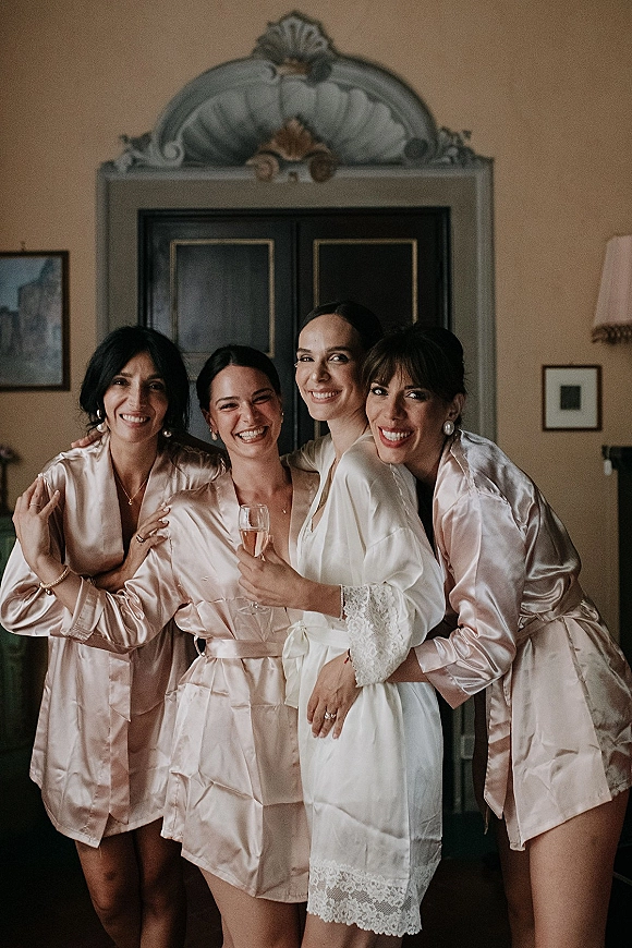 Bridesmaids getting ready in bridal party robes, smiling with champagne flutes in a bedroom near an ornate wardrobe and wall sconce