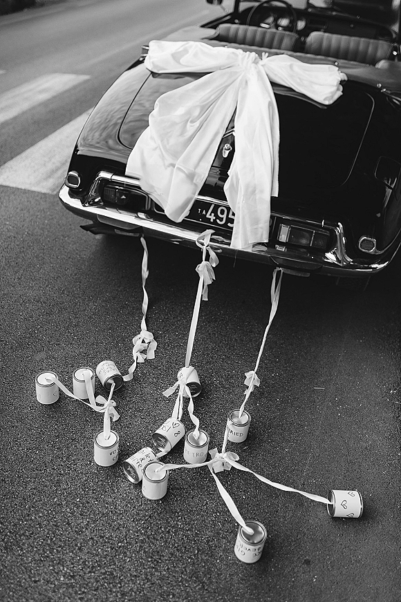 Wedding getaway car with an oversized bow, ribbons, and tin cans trailing behind a convertible on street pavement for the send-off