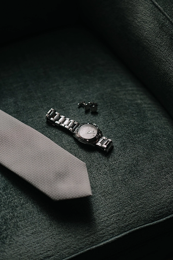 Groom accessories flatlay with groom watch detail, necktie and cufflinks arranged on textured upholstered chair fabric backdrop