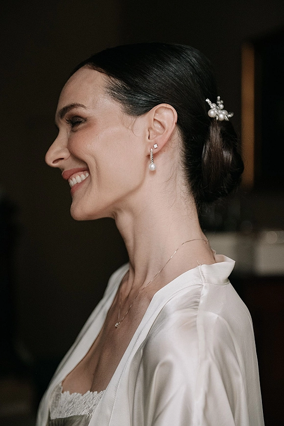 Bridal portrait of bride side profile in a silk bridal robe, smiling with pearl drop earrings and hair pins against a blurred indoor wall
