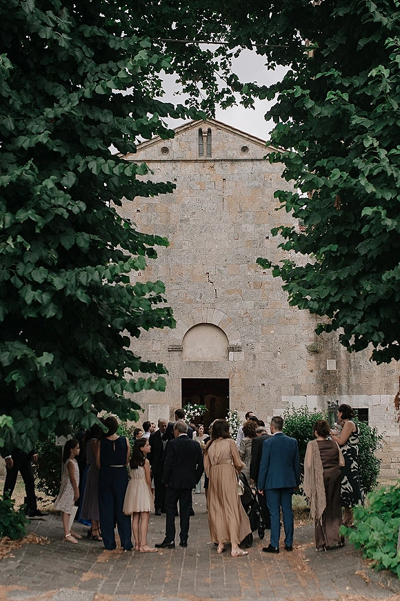 Wedding ceremony guests in suits and dresses gather at a church wedding entrance, passing floral arrangements and greenery by the stone doorway.