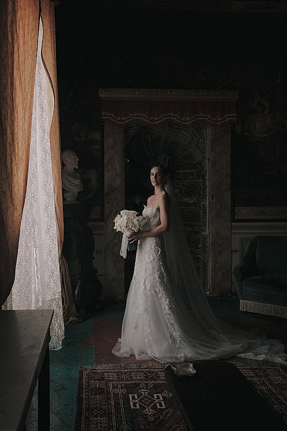 Bridal portrait of a bride in a strapless lace wedding dress with cathedral veil, holding a white bouquet in an ornate interior room