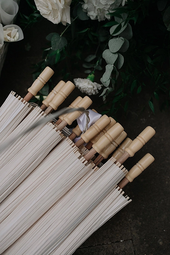 Wedding parasols with paper parasol wedding favors, white paper canopies with wooden handles, ribbon bow, and eucalyptus with white roses on dark stone pavement