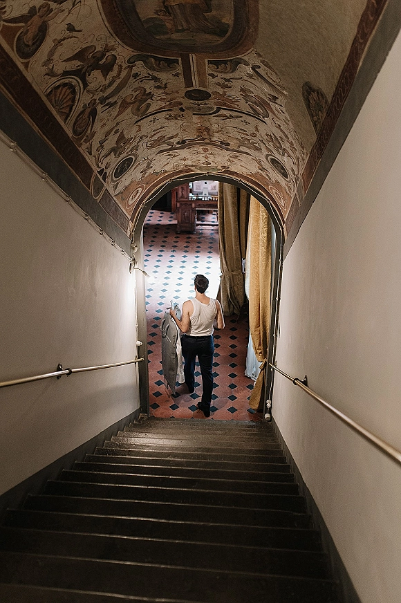 Groom getting ready, holding his suit jacket in an undershirt beside a staircase under an ornate fresco ceiling in a tiled hallway