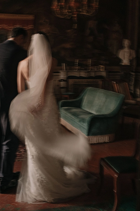 Wedding couple portrait of bride in strapless lace gown and veil with groom in dark suit beneath a chandelier in a moody lounge