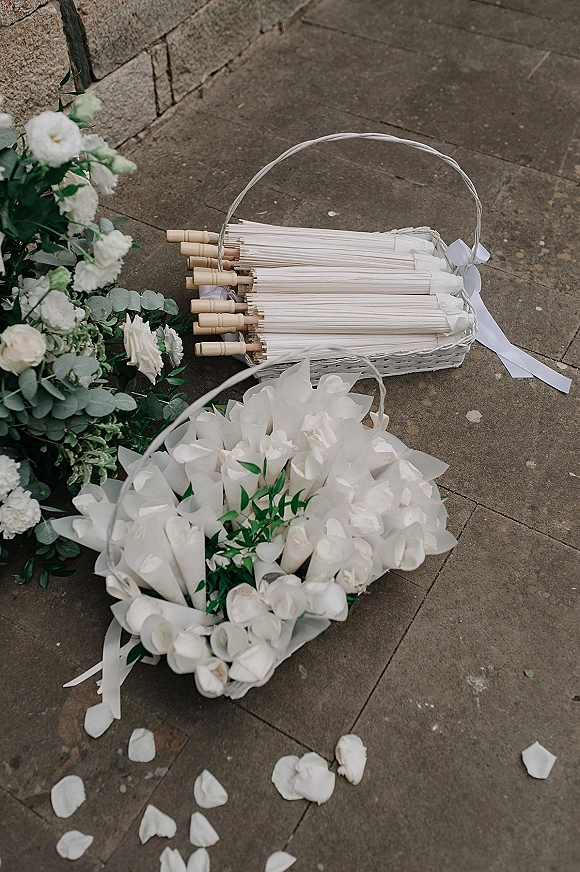 Wedding confetti cones in a wicker basket, paper confetti cones tied with white ribbon beside parasols on stone pavement