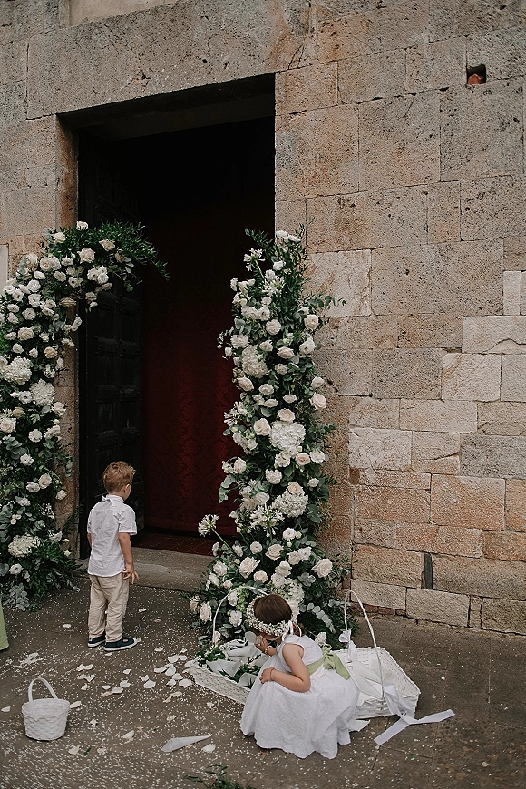 Flower girl and ring bearer holding a flower girl basket and ring pillow by a white floral arch at a stone doorway, petals underfoot
