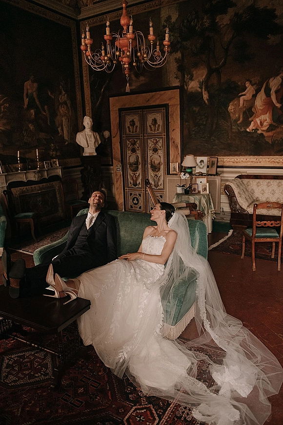 Couple portrait of bride and groom on sofa, laughing on a green velvet couch beneath a chandelier in an ornate vintage room