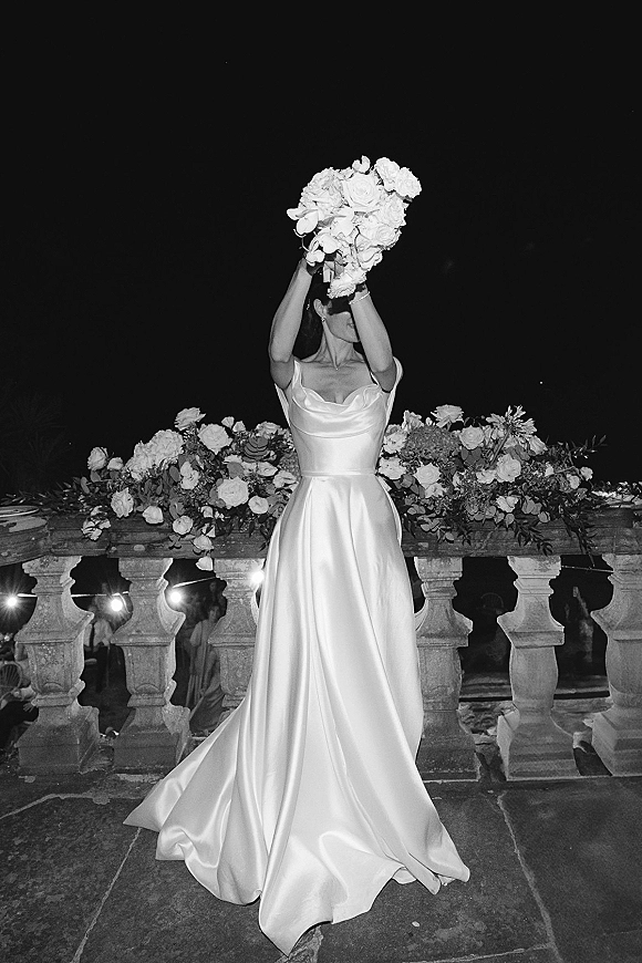 Bridal portrait of a bride lifting her bouquet overhead in a satin wedding dress on a terrace with stone balustrade and string lights at night