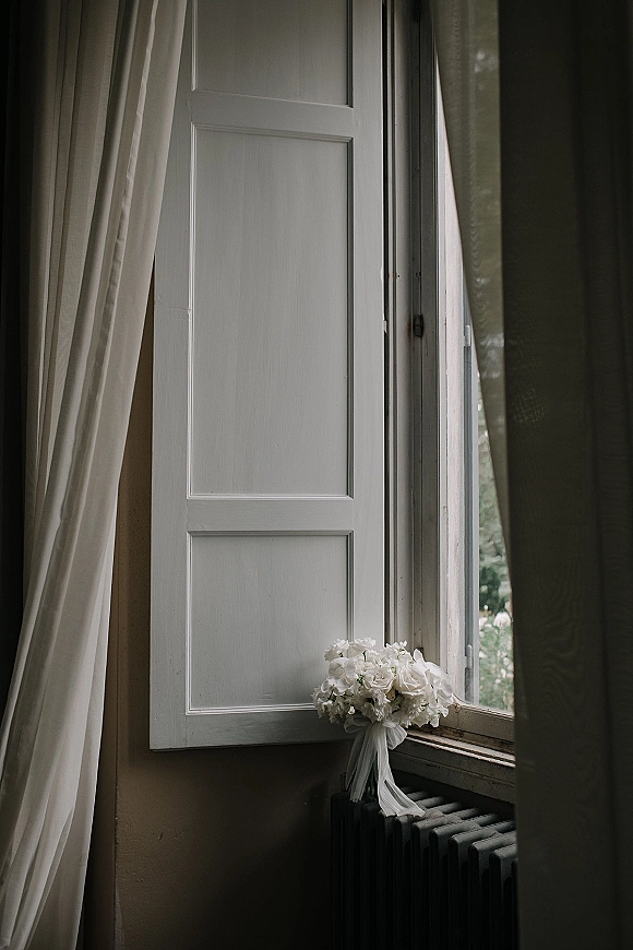 Bridal bouquet of white wedding bouquet flowers with a trailing ribbon on a windowsill by open shutters, soft window light and greenery outside