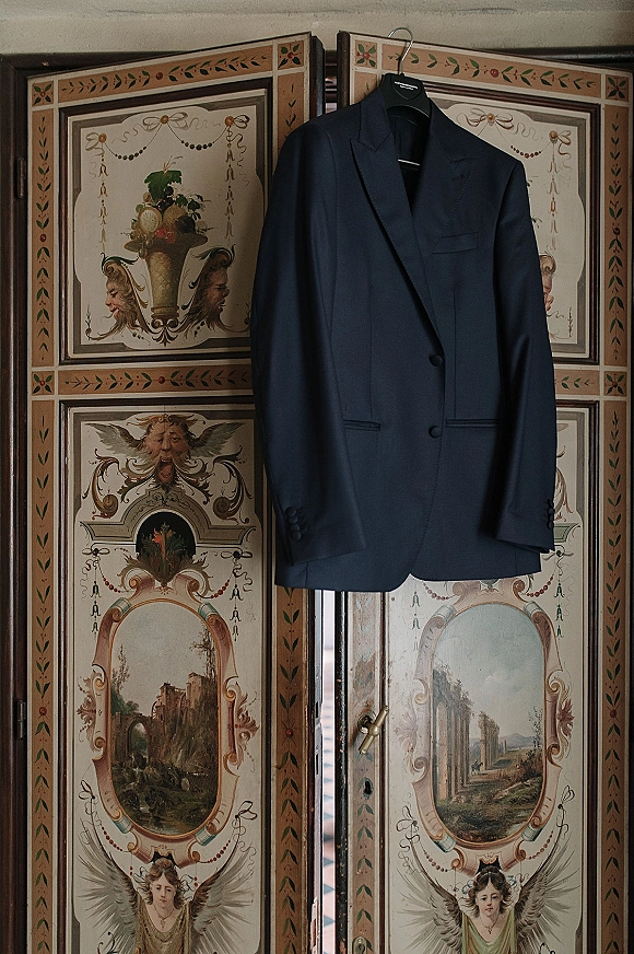 Groom suit jacket in a navy wedding suit on a hanger, highlighting buttons and lapels against a painted folding screen backdrop