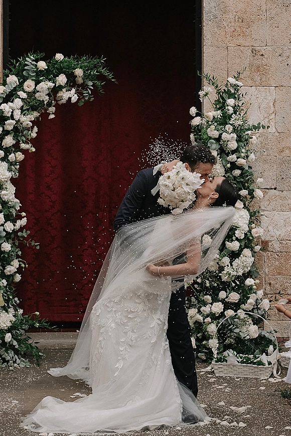 Wedding kiss as groom dips bride beneath a white floral arch, her long veil and bouquet framed by stone wall, red door, and confetti