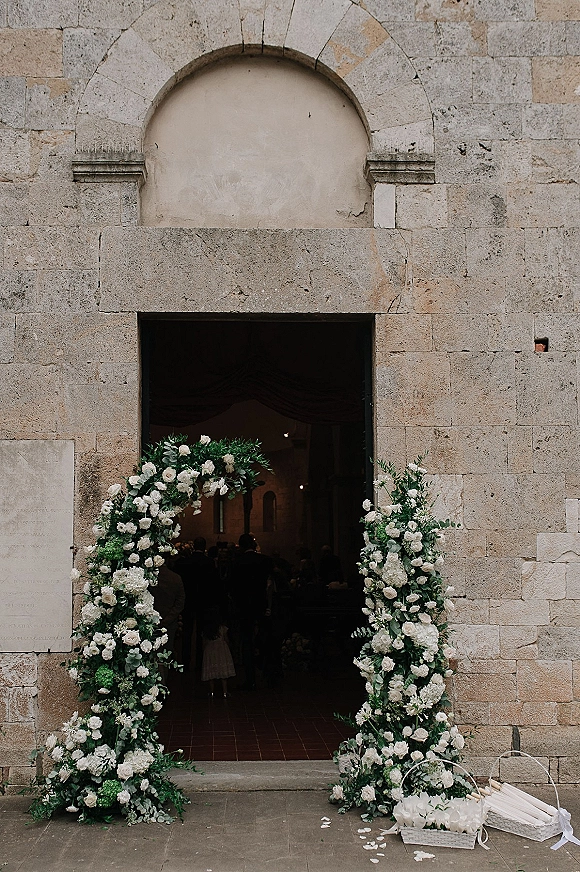 Wedding ceremony entrance with church entrance flowers framing a stone church doorway, white roses and greenery arch over petal-strewn aisle