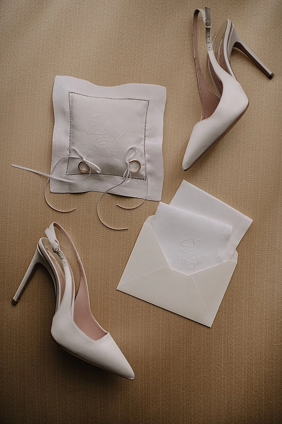Wedding details flat lay with wedding rings close up beside white slingback heels, monogrammed ring pillow and ribbon on linen backdrop