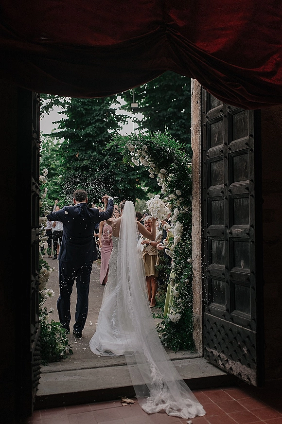 Wedding recessional as newlyweds walking out beneath a white-flower arch, bride’s veil trailing while guests toss confetti by wooden doors