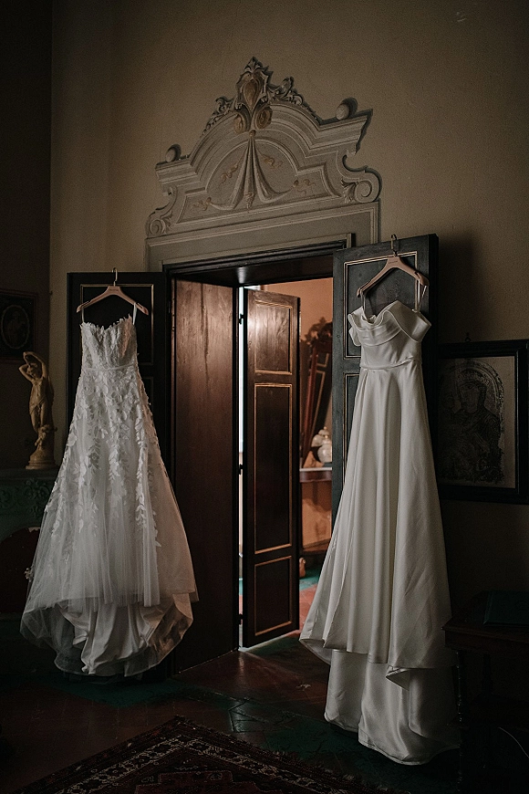 Wedding dresses hanging photo shows two gowns on wooden hangers in open dark wood doors, lace appliqué bodice and simple satin skirt in warm vintage room