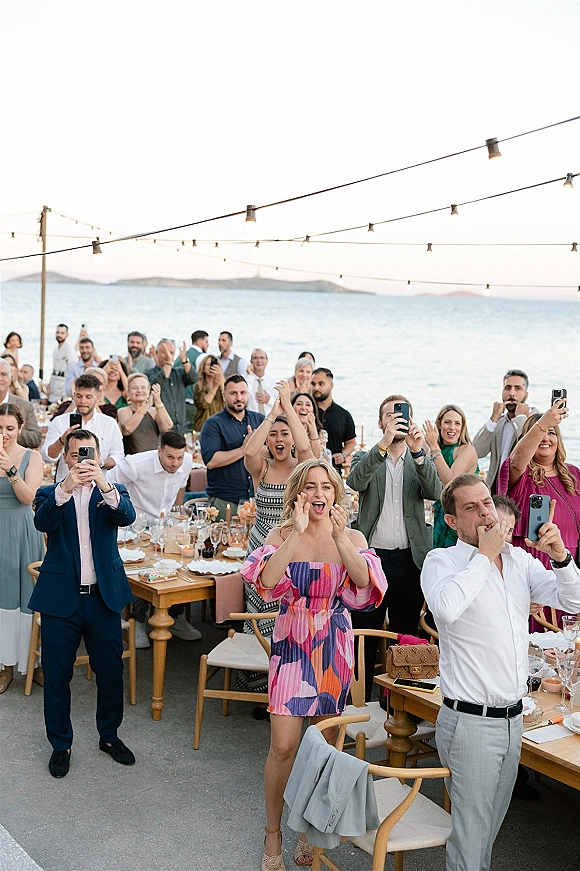 Wedding reception guests cheering and raising phones at long wooden tables under string lights, with candles and ocean horizon behind