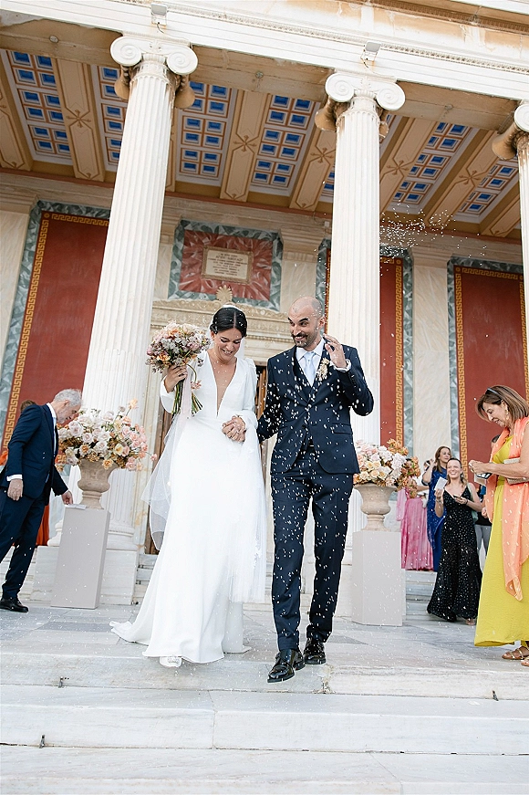 Wedding recessional as bride and groom walk out holding hands down stone steps, bride with bouquet and veil as guests throw confetti