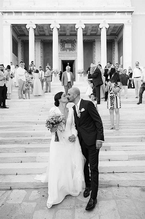 Wedding kiss portrait of newlyweds kissing on stone steps, bride with bouquet and veil, groom in suit, guests cheering by columns