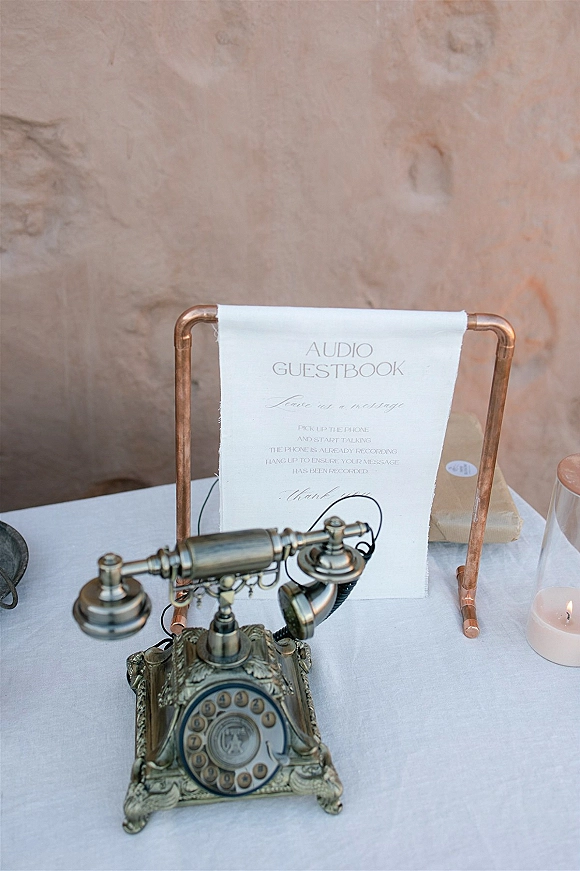 Wedding audio guestbook with an audio guestbook phone, vintage rotary telephone beside a sign on copper stand with candles by a stucco wall