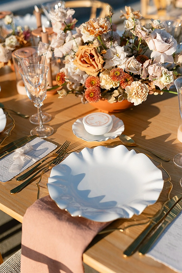 Reception tablescape with wedding table centerpiece in a terracotta bowl, roses and carnations, goblets, scalloped plates, gold flatware on wood table