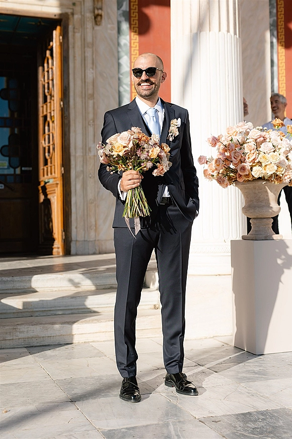 Groom portrait in a navy suit holding a bouquet, wearing sunglasses on marble steps by a columned entrance with floral pedestal arrangement