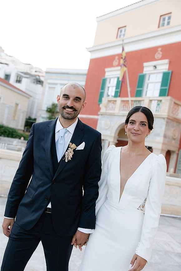 Couple portrait of bride and groom holding hands, smiling in a courtyard by a historic building with arched doorway and green shutters.