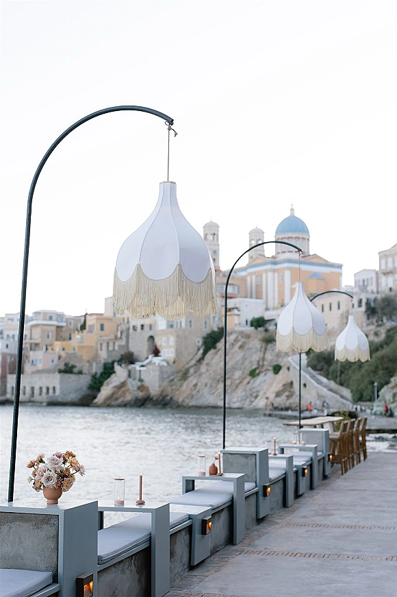Outdoor reception decor with waterfront wedding reception lounge seating, fringe pendant lamps, taper candles, and florals along a seaside promenade
