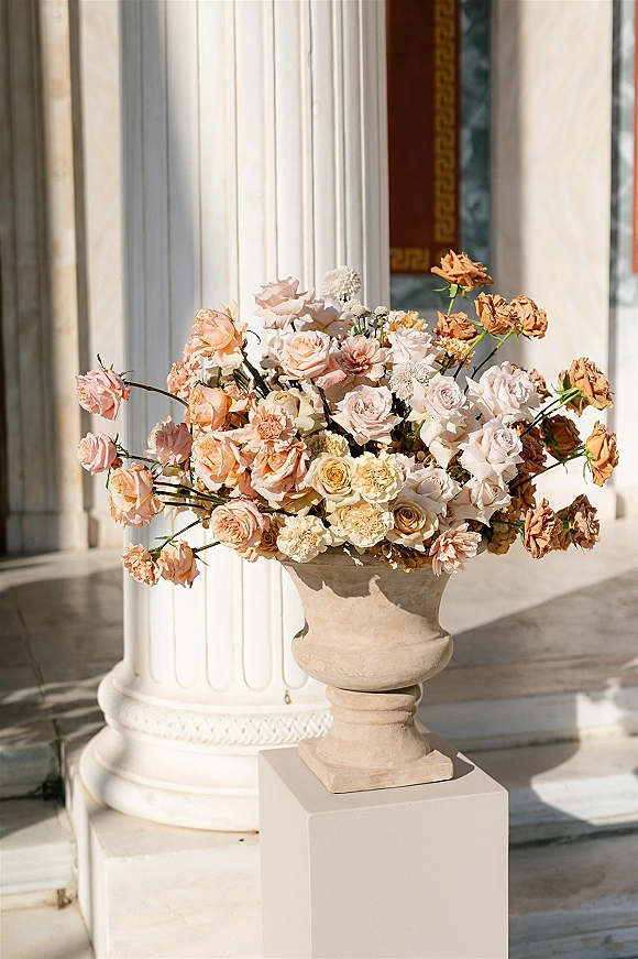 Wedding floral arrangement of peach and blush roses in a stone urn on a pedestal, set before white columns and stone steps