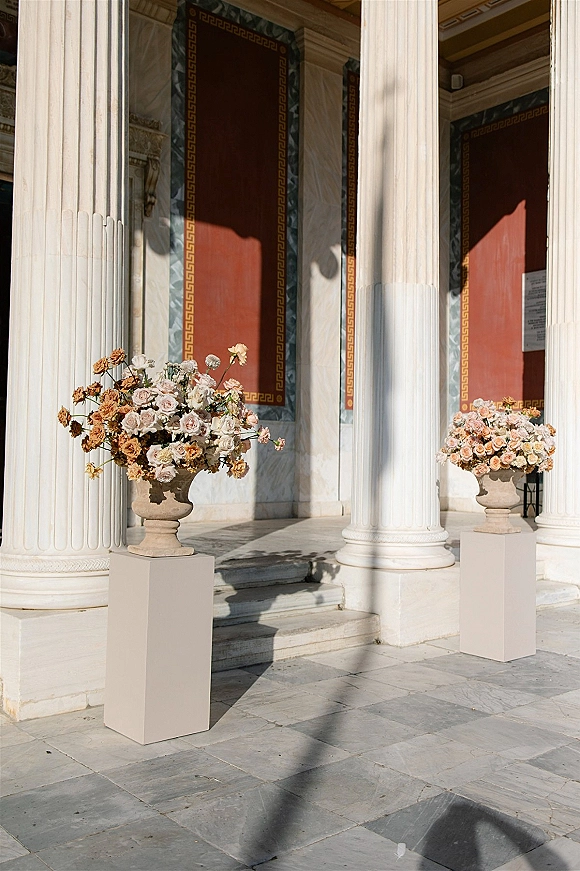 Wedding floral urns with ceremony entrance flowers in blush roses atop stone pedestals, flanking white columns and stone steps by red panels